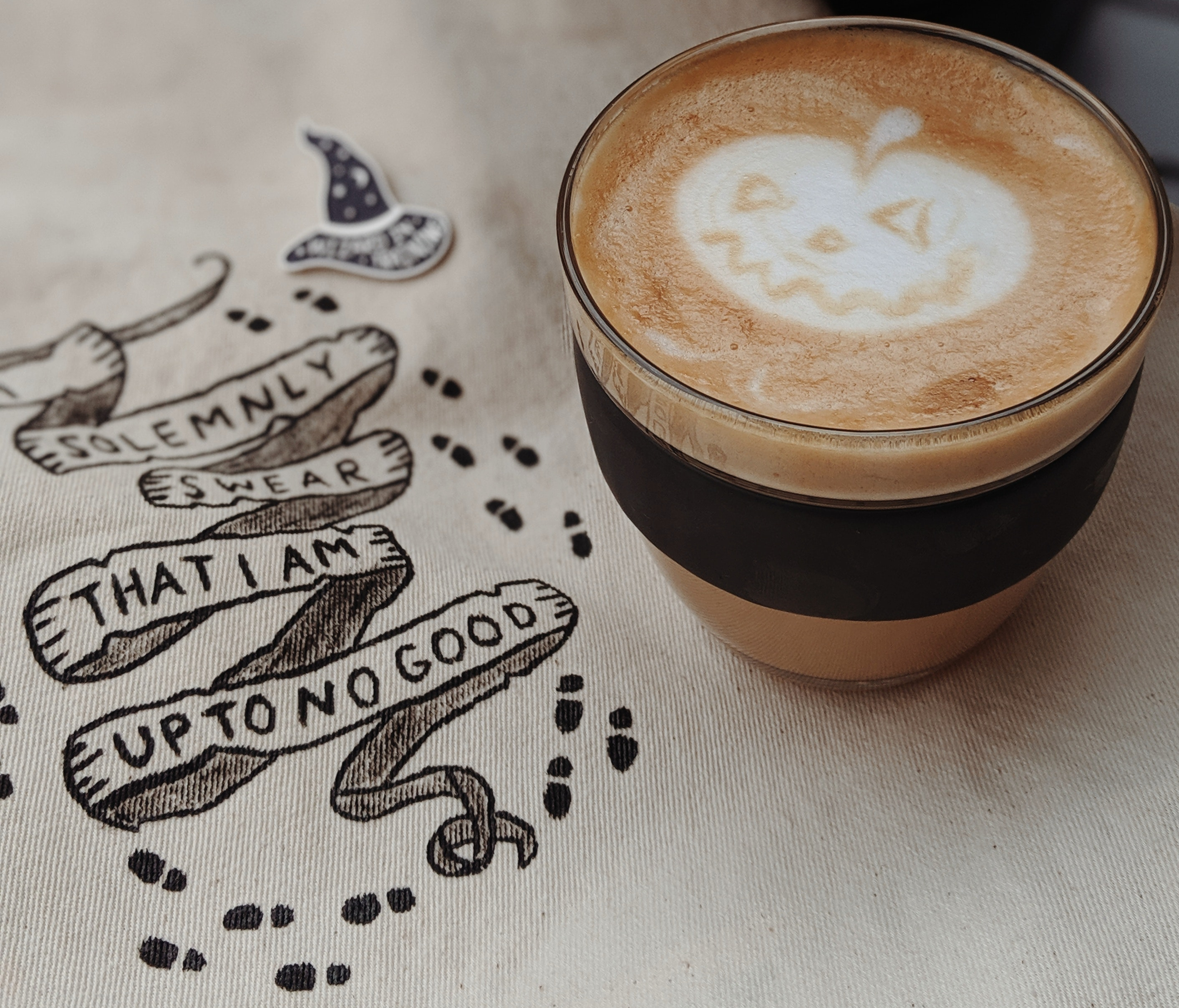 Coffee in glass mug onn fabric with print showing footstepps and a little magic hat reading: Solemnly swear that I am up to no good