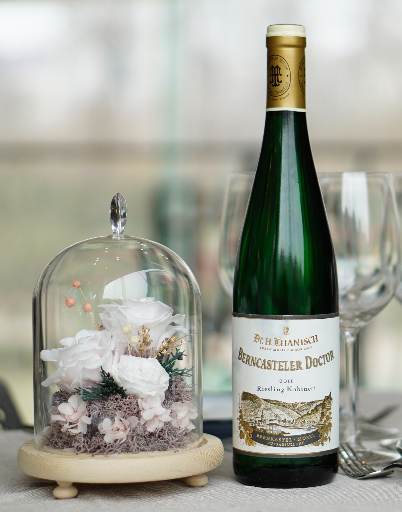 glass cloche with wooden base and white roses next to white wine bottle