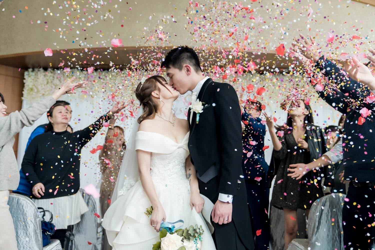 Kissing asian couple has mixed flower petal and paper confetti thrown at them by their guests